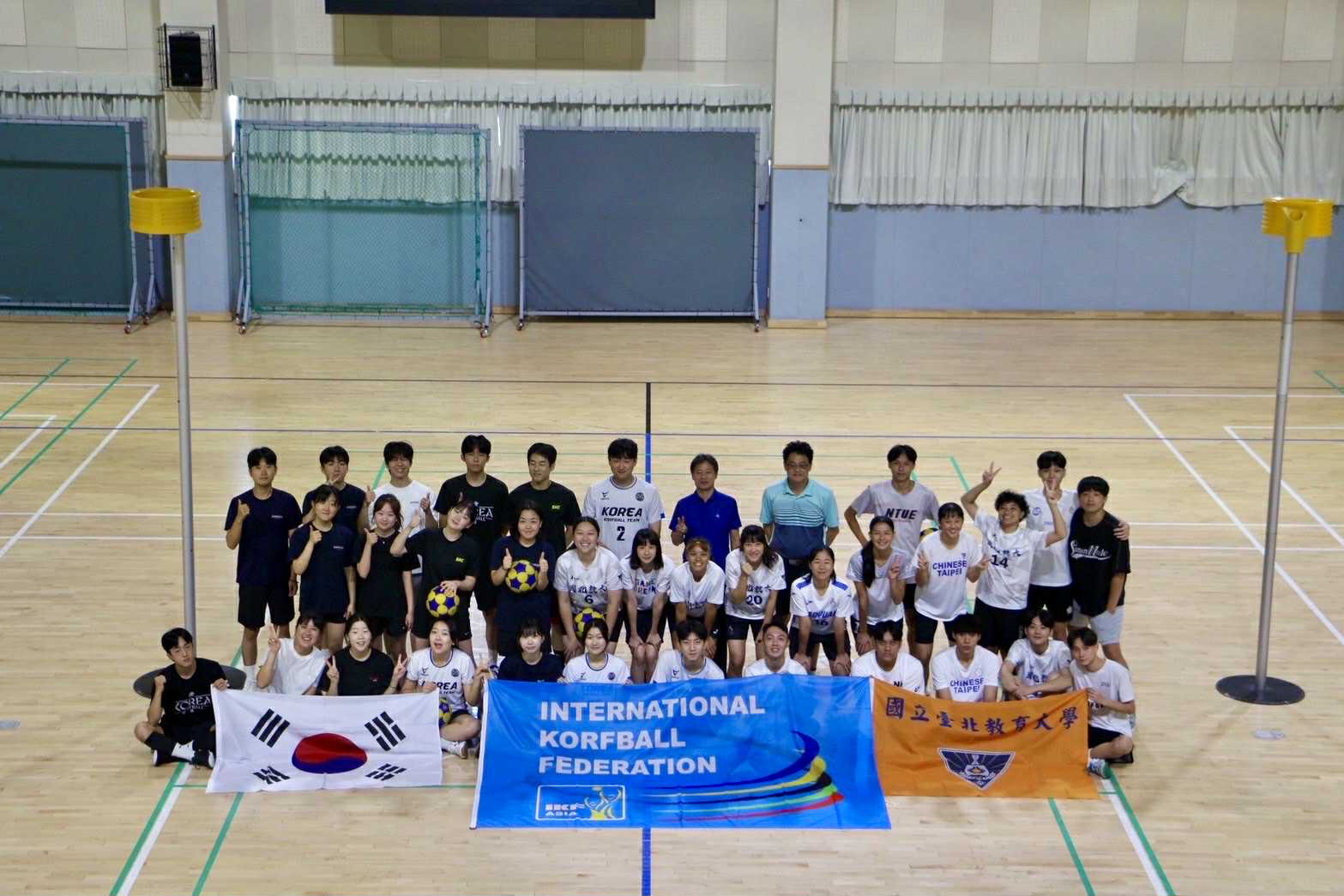  國立臺北教育大學與韓國的合球隊員，在室內合球場館拍攝團體大合照。眾人圍繞著一面「INTERNATIONAL KORFBALL FEDERATION」(國際合球總會) 的藍色會旗，並分別在兩側手持韓國國旗與「國立臺北教育大學」的橘色校旗，氣氛友好熱絡。