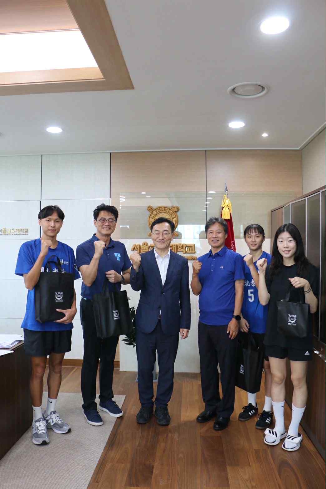 五位來自台灣的師生代表與一位穿著西裝的韓國校方人員在辦公室內合影。大家面帶微笑，一同舉起拳頭做出加油打氣的手勢。三位學生手上提著印有紀念標誌的黑色提袋，看起來是剛收到的贈禮。