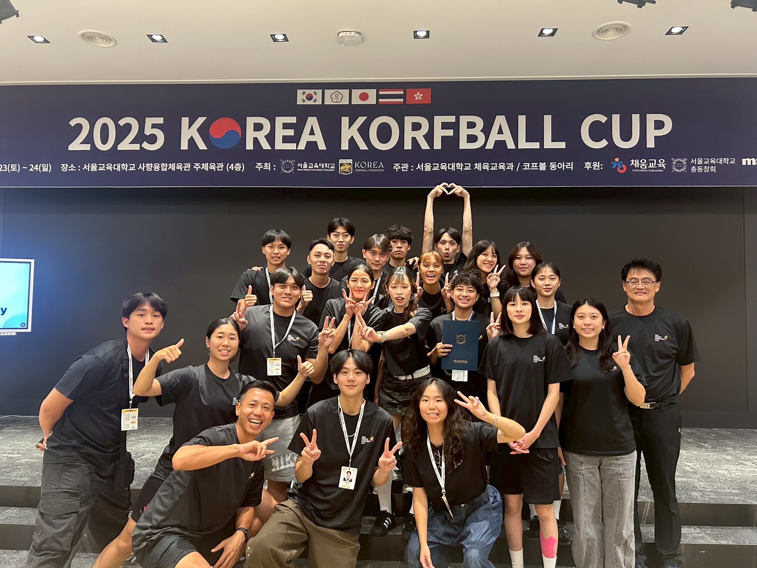 一支合球隊伍在「2025 KOREA KORFBALL CUP」藍色布條前拍攝團體照。隊員與教練們穿著黑色T恤，開心地對著鏡頭比出勝利手勢或愛心手勢，氣氛歡樂融洽。前排一位隊員拿著獎狀，布條上方有韓國、中華臺北、日本、泰國、與香港的旗幟。